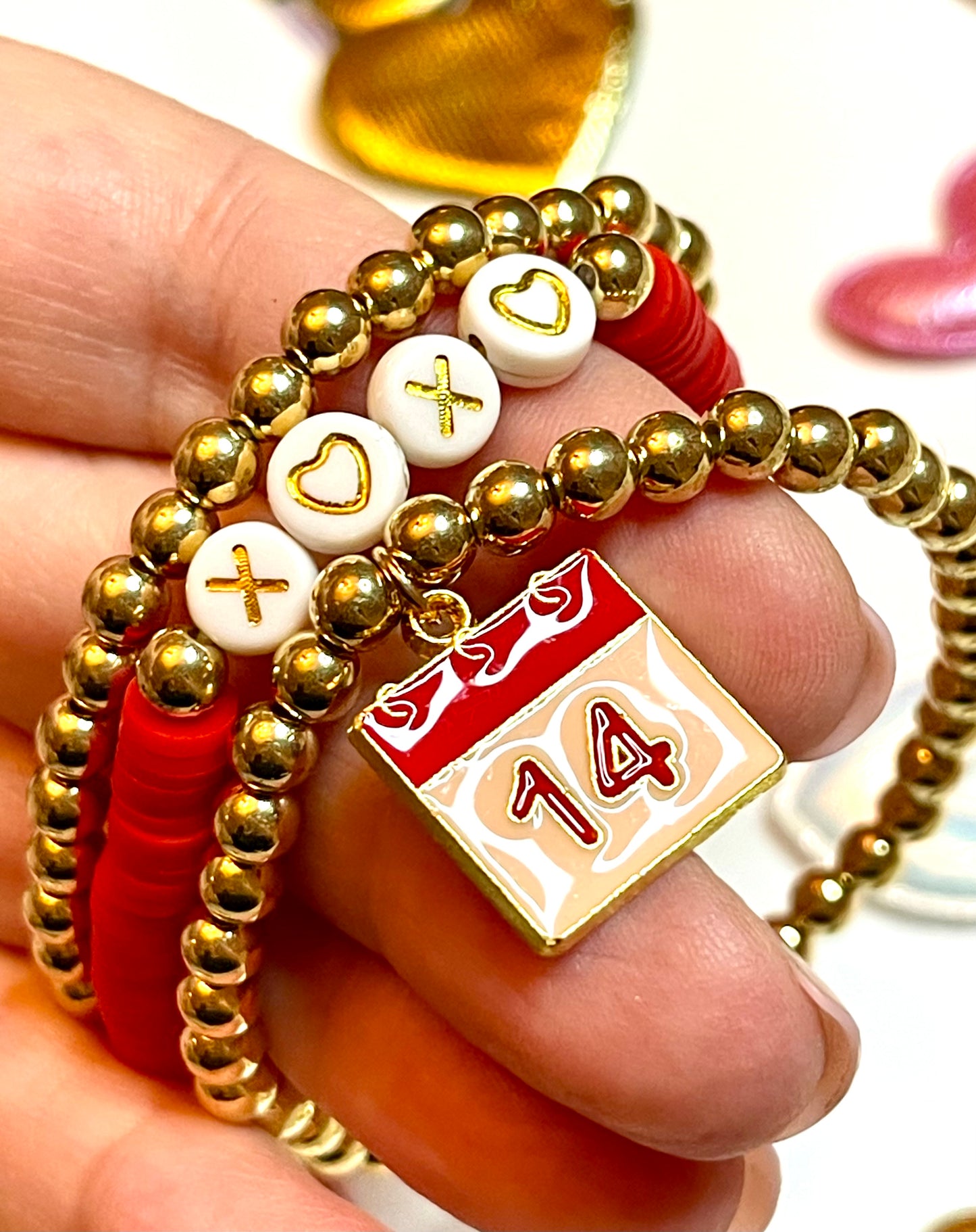 Love Stack 3: Two gold plated bead stretch bracelets, one with a 14th on the calendar charm and a red clay beaded bracelet with letter beads